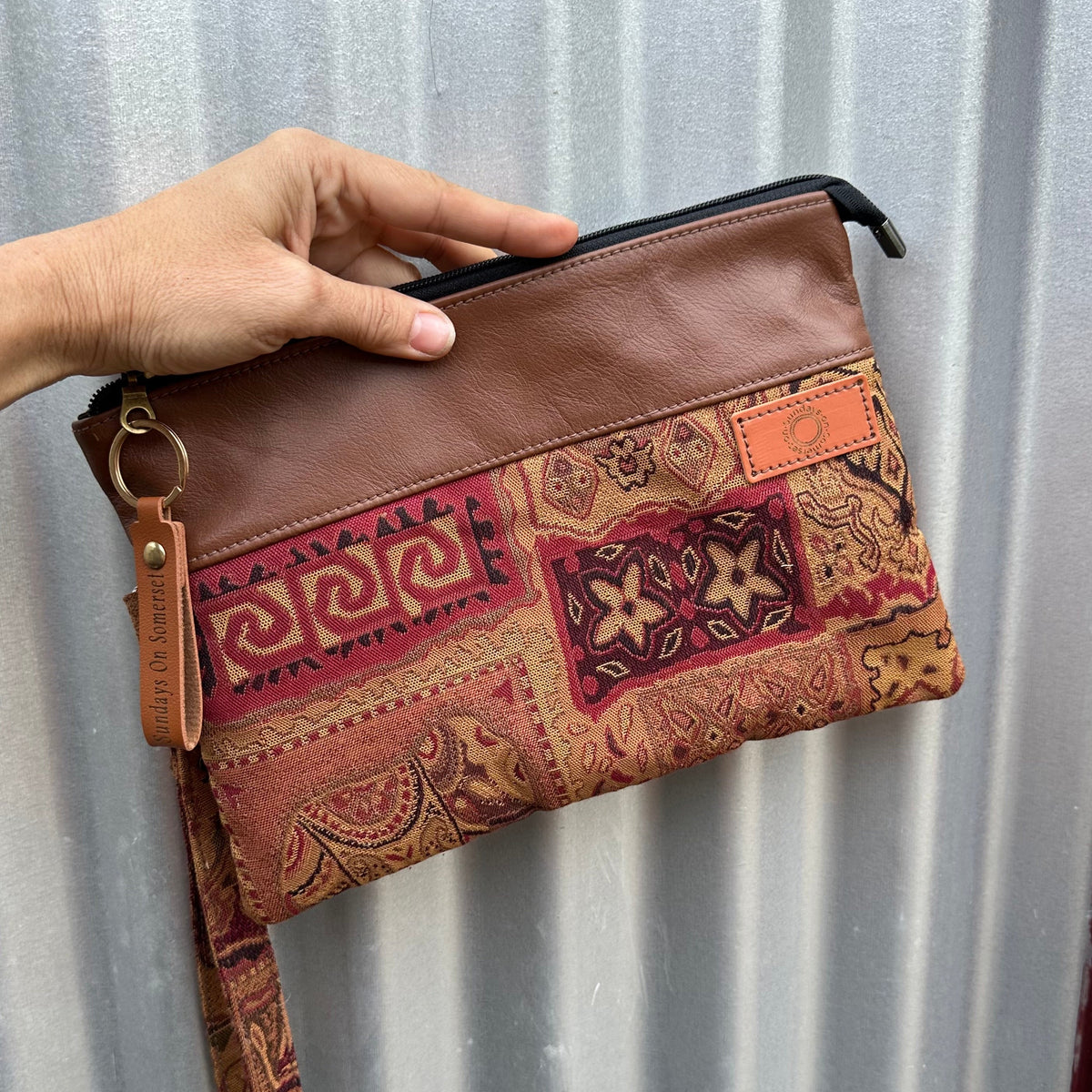 A hand holds a brown leather zip pouch with a bohemian patchwork fabric sleeve featuring red, orange, and brown patterns, set against a corrugated metal background.