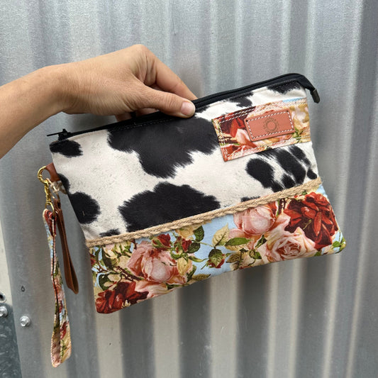 A wristlet clutch zippered pouch featuring a cow-print top section, a floral rose pattern bottom section, and a patch on the front