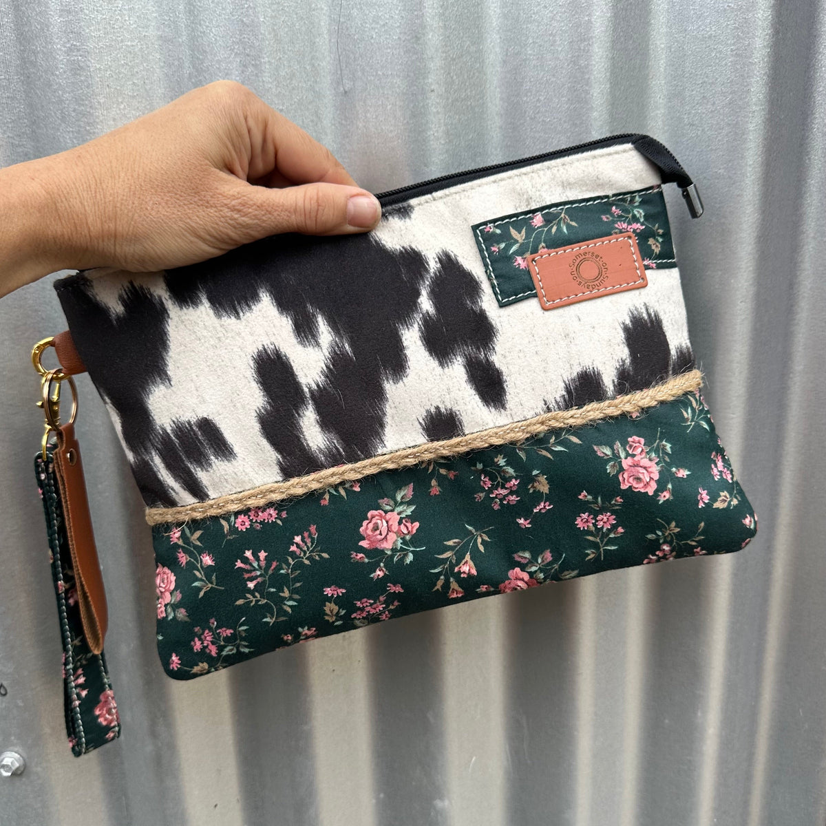 A clutch wristlet zip pouch with a cow-print top half, a dark green floral bottom half, and a  patch on the front,