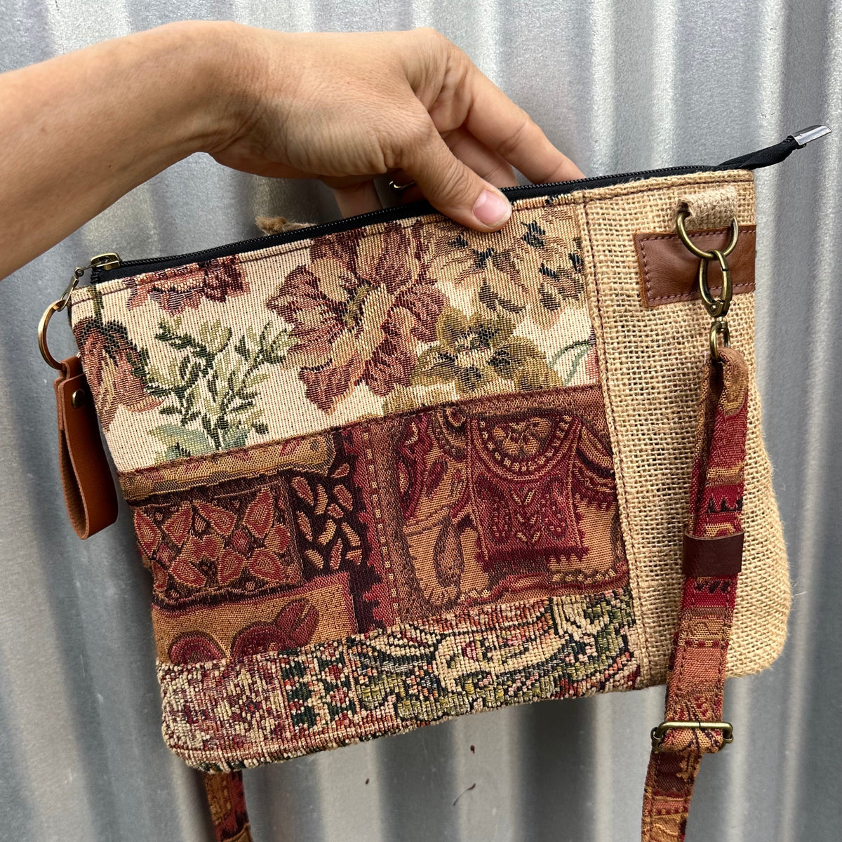 A hand holds a small patchwork fabric clutch bag with a floral and paisley pattern, brown leather trim, and a matching shoulder strap against a corrugated metal background.