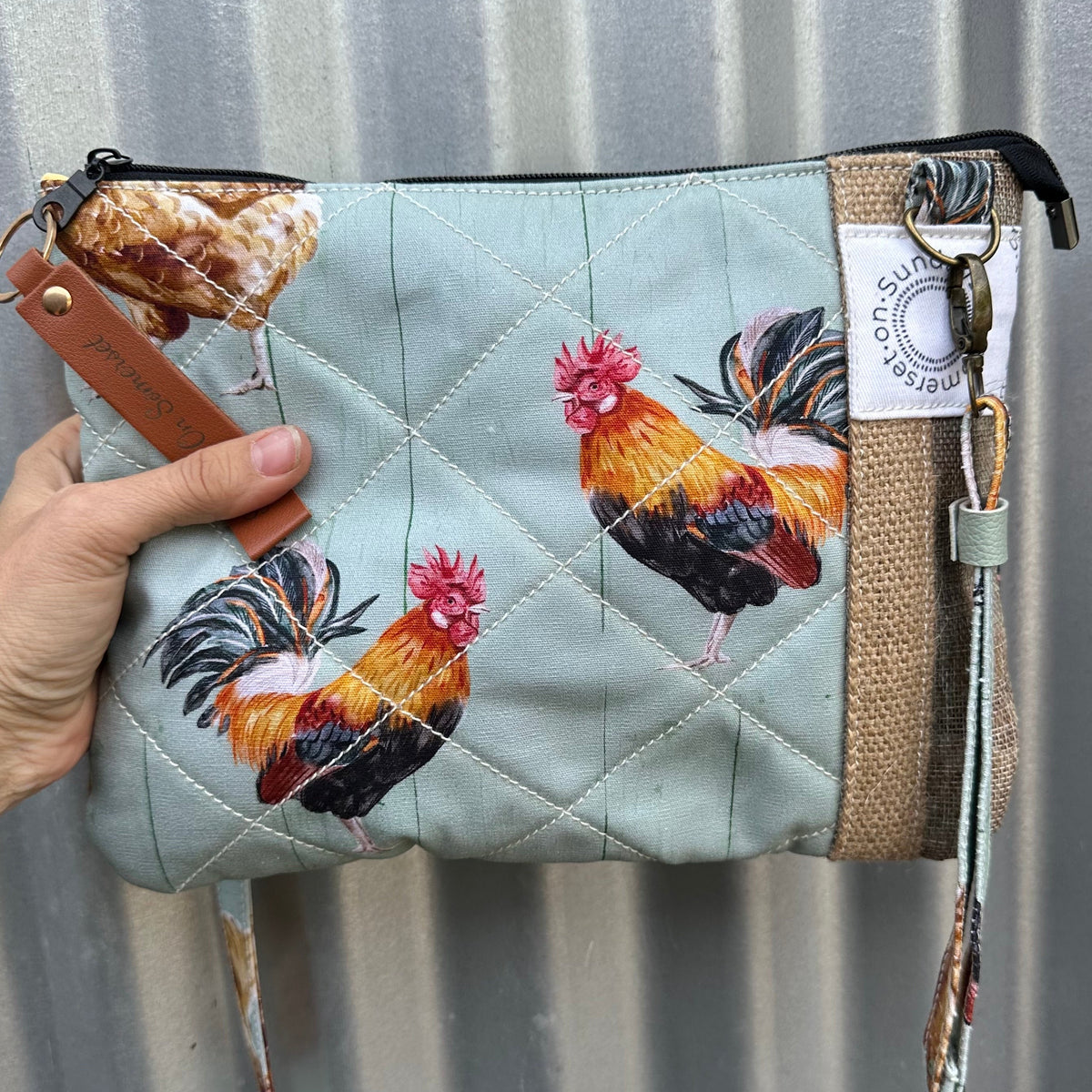 A quilted clutch bag in a pale blue-green fabric with printed rooster and chicken illustrations, held by a hand against a corrugated metal background. The bag features a tan burlap-textured side panel, a wristlet strap, and a white brand tag reading 'Sundays on Spmerset'.