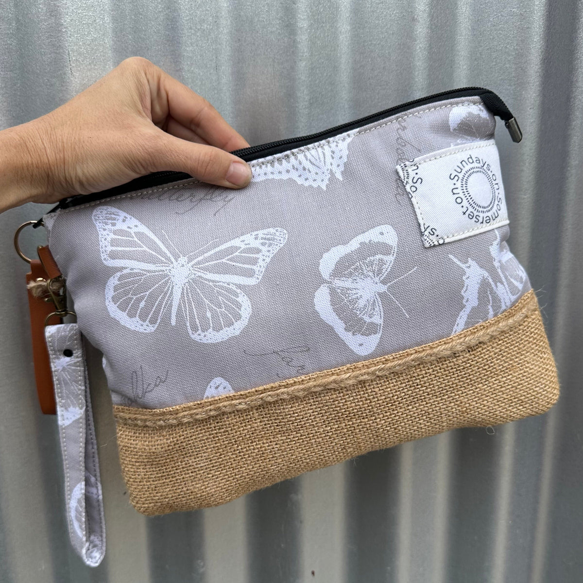 A hand holds a zippered pouch with a light gray butterfly print and a natural burlap bottom, against a corrugated metal background.