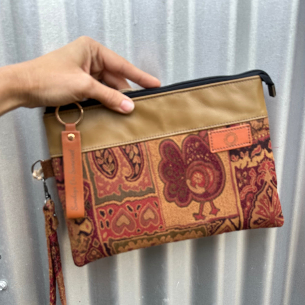 A hand holds a zip pouch with a tan top panel and a multicolor paisley-patterned lower section, featuring a wrist strap and key ring against a corrugated metal background.
